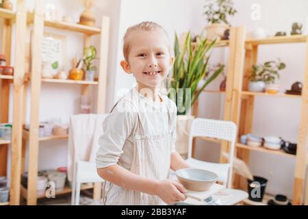 Portrait d'un jeune garçon gai dans un tablier avec une plaque d'argile dans ses mains, dans le studio de poterie. Image dans des tons chauds et ensoleillés Banque D'Images