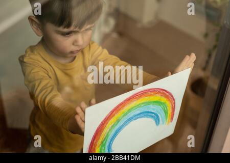 Un petit enfant inspirant tenant un dessin d'un arc-en-ciel à travers la fenêtre Banque D'Images