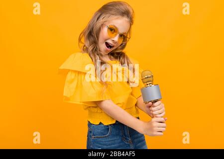 Portrait de la jeune fille caucaienne avec de longs cheveux équitables dans des verres jaunes sourires et chante des chansons avec un microphone Banque D'Images