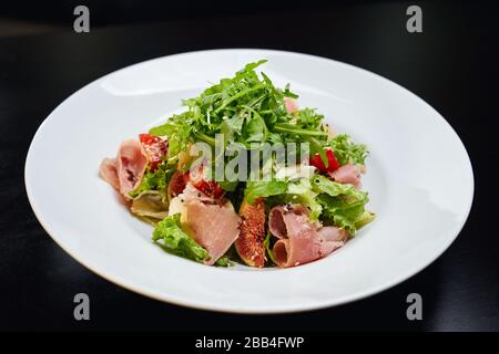 Salade délicieuse avec jambon, légumes frais, figues, laitue et roquette à salade sur le dessus. Vue de dessus d'un plat frais et sain servi avec de la sauce brune et du sésame sur une assiette blanche au restaurant. Banque D'Images