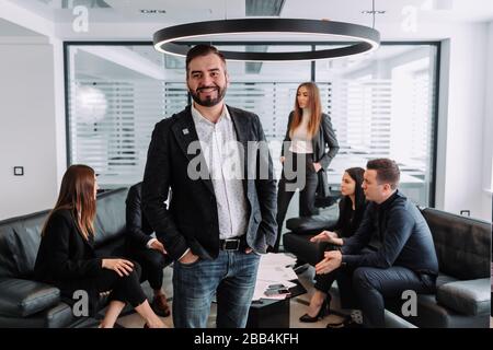 Homme d'affaires confiant. Un jeune homme beau regarde la caméra alors que ses collègues discutent de quelque chose en arrière-plan Banque D'Images