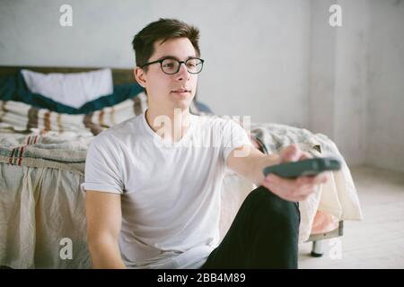 Guy utilisant la télécommande tout en étant assis sur le lit à la maison. Joyeux jeune homme qui regarde la télévision . Émotions humaines sur son visage. Banque D'Images