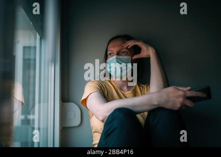 Femme en auto-isolation lors d'une épidémie de virus à l'aide d'un téléphone portable. Femme inquiète avec masque chirurgical de protection, tenant smartphone, sélectif f Banque D'Images