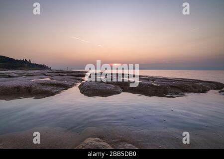 plage de sirmione la nuit et coucher de soleil sur le lac de Garde avec rochers plats sur la plage et l'eau et le soleil dedans l'horizon Banque D'Images