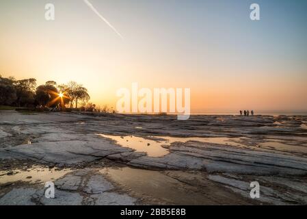 plage de sirmione la nuit et coucher de soleil sur le lac de Garde avec rochers plats sur la plage et l'eau et le soleil dedans l'horizon Banque D'Images