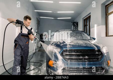 Tir de jeune homme caucasien laver la voiture bleue sous l'eau haute pression dans le service de lavage de voiture. Retrait du savon avec de l'eau, nettoyage de voiture Banque D'Images