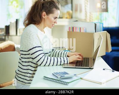 femme d'affaires moderne en âge moyen dans un pull rayé dans un bureau à domicile temporaire dans la maison moderne en journée de travail ensoleillée. Banque D'Images