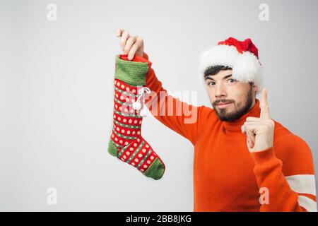 Un jeune homme barbu isolé sur fond. Un gars sérieux et confiant présente une chaussette dans la main. Pointez vers le haut avec le doigt. Portez un caillot rouge de Noël ou de nouvel an Banque D'Images