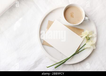 Petit déjeuner dans la scène de maquette près de la fenêtre. Tasse de cercueil, carte de vœux, narcisse, fleurs de jonquille sur plateau en marbre fond de toile de table en lin blanc Banque D'Images