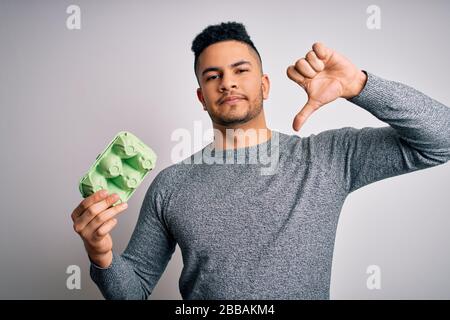 Jeune homme de handsome tenant carton boîte d'œufs frais sains sur fond blanc avec visage en colère, signe négatif montrant déaimer avec les pouces vers le bas, rej Banque D'Images