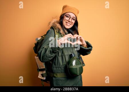 Jeune femme randonneur portant un sac à dos, une cantine et une carte sur fond jaune souriant en amour montrant le symbole du coeur et la forme avec les mains. Romantique Banque D'Images