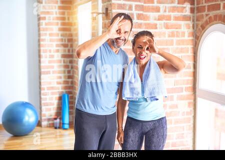 Couple sportif d'âge moyen debout avant de faire de l'exercice à la salle de gym faire un geste ok avec le sourire de la main, l'oeil regardant par les doigts avec le visage heureux. Banque D'Images