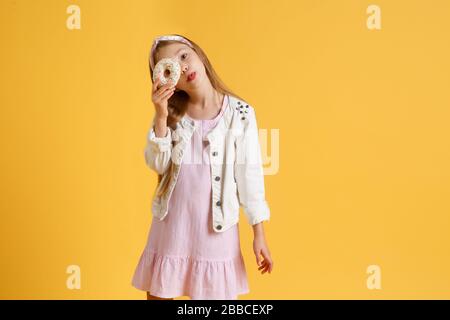 Une jeune fille blonde folle et gaie dans un chapeau d'anniversaire souriant, s'amuser et regarder par deux beignets rouges sur ses yeux. Bonbons. Fond de studio jaune. Banque D'Images