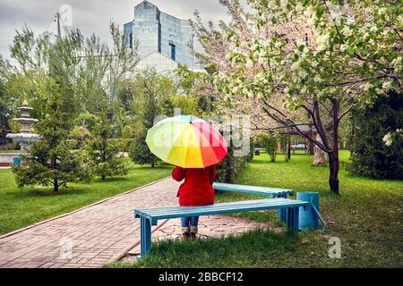 Femme en robe rouge avec parapluie arc-en-ciel assis dans le parc avec arbres en fleurs Banque D'Images