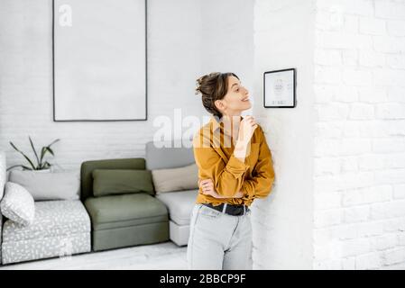 Jeune femme contrôlant la température dans le salon avec un écran tactile numérique. Concept de contrôle du chauffage dans une maison intelligente Banque D'Images