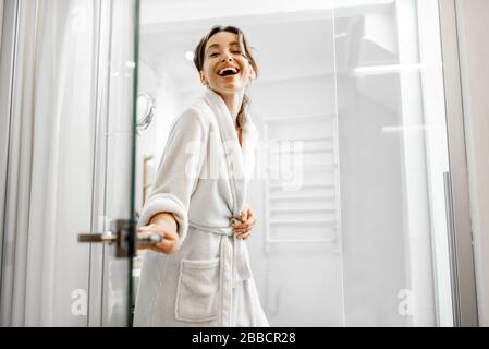 Jeune et joyeuse femme en peignoir allant à la salle de bain, fermant une porte en verre à la maison Banque D'Images