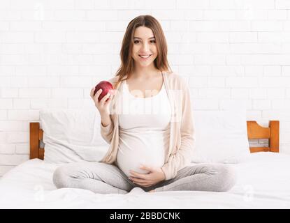 Jeune femme enceinte mangeant de la pomme rouge dans le lit à la maison Banque D'Images