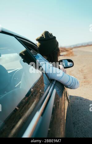 une jolie fille brunette dans un chapeau tricoté militaire et un sweat à capuche regarde devant le ciel bleu clair dans la fenêtre ouverte d'une voiture vert foncé Banque D'Images