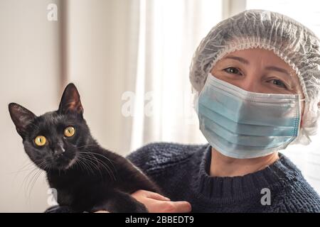 Une femme dans un masque pour se protéger contre le coronavirus, tenant un chat noir dans ses bras Banque D'Images