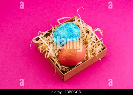 L'oeuf de Pâques est de couleur bleue avec or et orange avec or. Deux œufs se trouvent dans une boîte de copeaux. Oeuf de pâques peint sur un fond rose. Espace de copie Banque D'Images