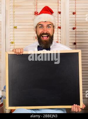 Père Noël dans un chapeau rouge avec un visage gai. L'homme avec barbe et noeud papillon tient un tableau noir blanc, espace de copie. Concept de célébration et d'humeur du nouvel an. Guy avec tableau de surveillance sur fond de mur en bois. Banque D'Images