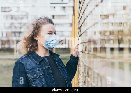 Une femme concernée d'Europe de l'est portant un masque médical à l'extérieur, se tenant à côté de la clôture en mesh avec un bâtiment résidentiel en arrière-plan. Virus Corona Banque D'Images