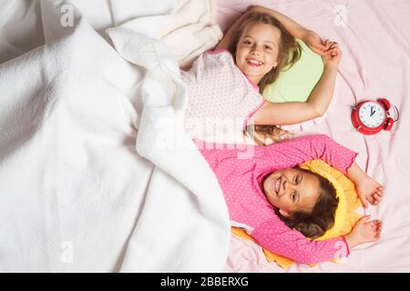 Écolières ayant une soirée de pyjama. Les enfants avec des visages souriants se trouvent sur un fond de couverture rose et blanc, vue de dessus. Enfants en pyjama rose près de l'horloge rouge. Concept d'enfance, de bonne matinée et de bonheur Banque D'Images