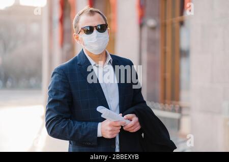 Virus, pandémie, concept de travail. Le Manager masculin porte un costume formel et des lunettes de soleil, un masque médical de protection pour prévenir le coronavirus, tient des papiers dans les mains, p Banque D'Images