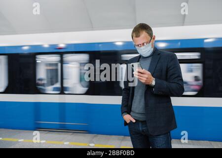 Le passager masculin porte des masques poses sur la plate-forme, attend le train, se rendre dans le métro, concentré dans le smartphone, lit les nouvelles en ligne. Viru Banque D'Images
