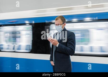 Jeune homme Manager utilise le téléphone portable, empêche la propagation de Coronavirus, pose contre le métro, pose à la plate-forme, vérifie les nouvelles en ligne, porte médical Banque D'Images