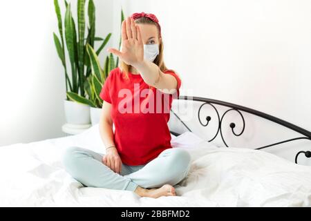 Une jeune blonde est assise sur un lit blanc pendant la période de quarantaine à cause du virus COVID-19. Elle porte un T-shirt rouge, confortable h Banque D'Images