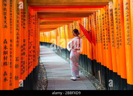 Jeune femme japonaise regardant derrière porter une robe orientale avec un parapluie marchant à l'intérieur des portes torii au sanctuaire de Fushimi Inari à Kyoto, au Japon. Banque D'Images