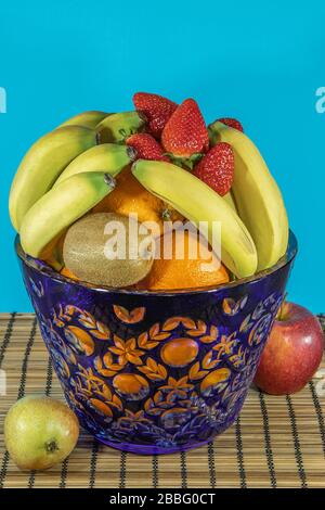 Fruits tropicaux et subtropicaux frais dans un grand vase en cristal bleu. Support en osier en bambou sur la table à manger, fond bleu. Banque D'Images