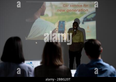 Un développeur d'applications créatives afro-américain et joyeux, se tenant contre l'écran de projection et présentant une idée pour le service de voiture aux investisseurs Banque D'Images