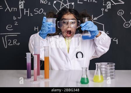 drôle de scientifique enfant avec des gants et des lunettes de protection en blouse de laboratoire avec des fioles chimiques, fond de tableau noir avec des formules scientifiques, explosion dans le labourato Banque D'Images