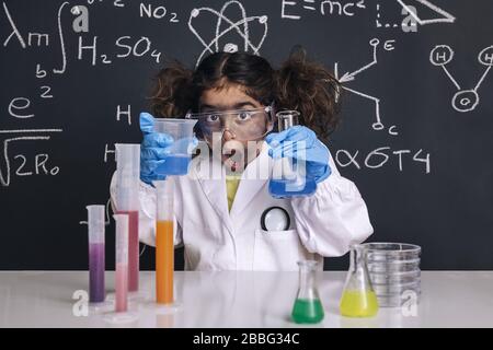 surprise scientifique fille avec des gants et des lunettes de protection dans un manteau de laboratoire avec des fioles chimiques, fond de tableau noir avec des formules scientifiques, explosion dans le travail Banque D'Images