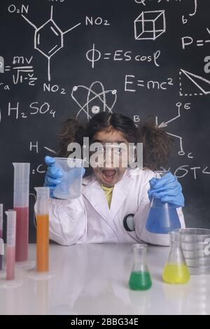 une fille scientifique folle avec des gants et des lunettes de protection en blouse de laboratoire avec fioles chimiques, fond de tableau noir avec des formules scientifiques, explosion dans l'ouvrier Banque D'Images