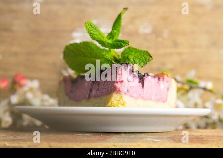 Des tranches de mousse de myrtilles avec des baies décorées de verglas miroir et des feuilles de menthe sur une plaque blanche avec des fleurs blanches sont sur table en bois, à proximité Banque D'Images