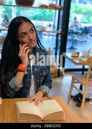 Jolie fille urbaine assise dans un café, lecture d'un livre et parler sur un téléphone mobile , Panama, Amérique centrale Banque D'Images