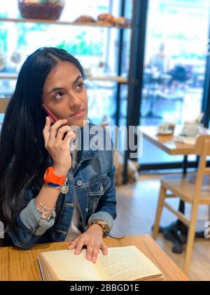 Jolie fille urbaine assise dans un café, lecture d'un livre et parler sur un téléphone mobile , Panama, Amérique centrale Banque D'Images