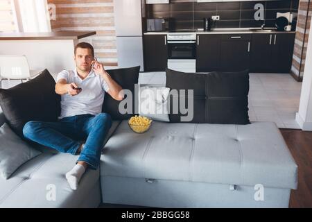 Jeune homme regarder la télévision dans son propre appartement. Un gars occupé assis sur le canapé et parler au téléphone. Changement de chaînes tv avec télécommande. Divertissez-vous Banque D'Images