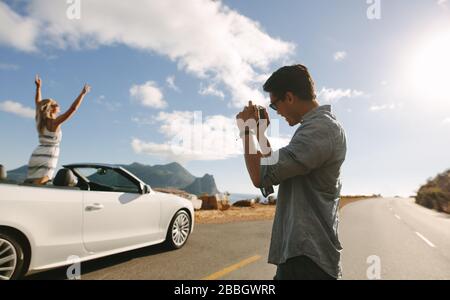 Homme avec appareil photo prendre des photos d'une femme debout dans un cabriolet. Femme debout avec ses mains soulevées à l'intérieur d'une voiture avec son petit ami prenant son pH Banque D'Images