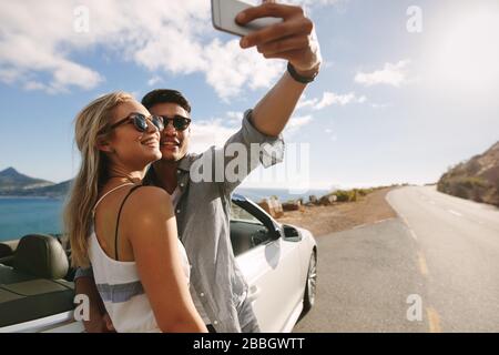 Bel homme et femme portant des lunettes de soleil prenant la photo selfie ensemble tout en se tenant en voiture sur la route. Banque D'Images