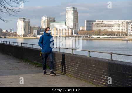 Le mâle du milieu des années 40 porte une veste bleue et un masque de visage marche le long de la Tamise, avec le quartier financier de Londres et Canary Wharf en arrière-plan. Banque D'Images