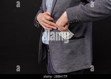 Un homme dans une veste met des dollars dans sa poche sur un fond noir. Gros plan. Banque D'Images