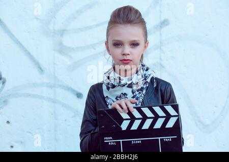 Charmante petite fille heureuse dans une jolie veste en cuir noir et un foulard noir blanc tient un clapper de film, regardant l'appareil photo sérieux, debout sur blanc Banque D'Images