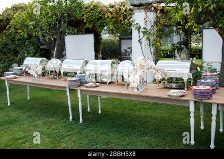 Soirée extérieure de printemps ou d'été dans un jardin décontracté pour le dîner-déjeuner avec table longue, fleurs, assiettes et nappe blanche Banque D'Images