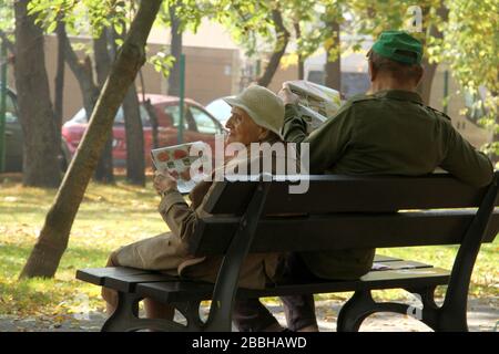 Bucarest, Roumanie. Femme âgée avec l'épicerie hebdomadaire et homme lisant le journal sur un banc de ville. Banque D'Images