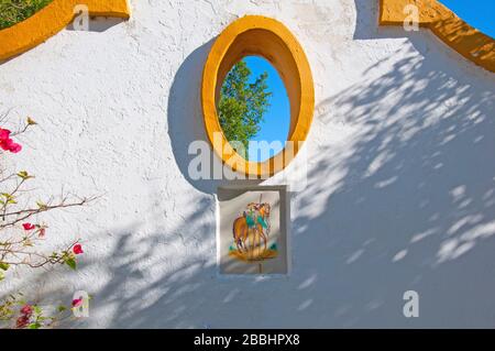 Mur blanc avec décorations orange, fenêtre ovale et carrelage espagnol. Buisson avec fleurs rouges, journée ensoleillée à Isla Moyor, Séville, Espagne Banque D'Images