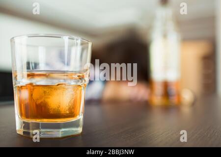 Jeune femme dans la cuisine pendant la quarantaine. Verre avec whisky ou une autre boisson alcoolisée devant. Sleppy ivre femme avec bouteille sur fond flou. Banque D'Images